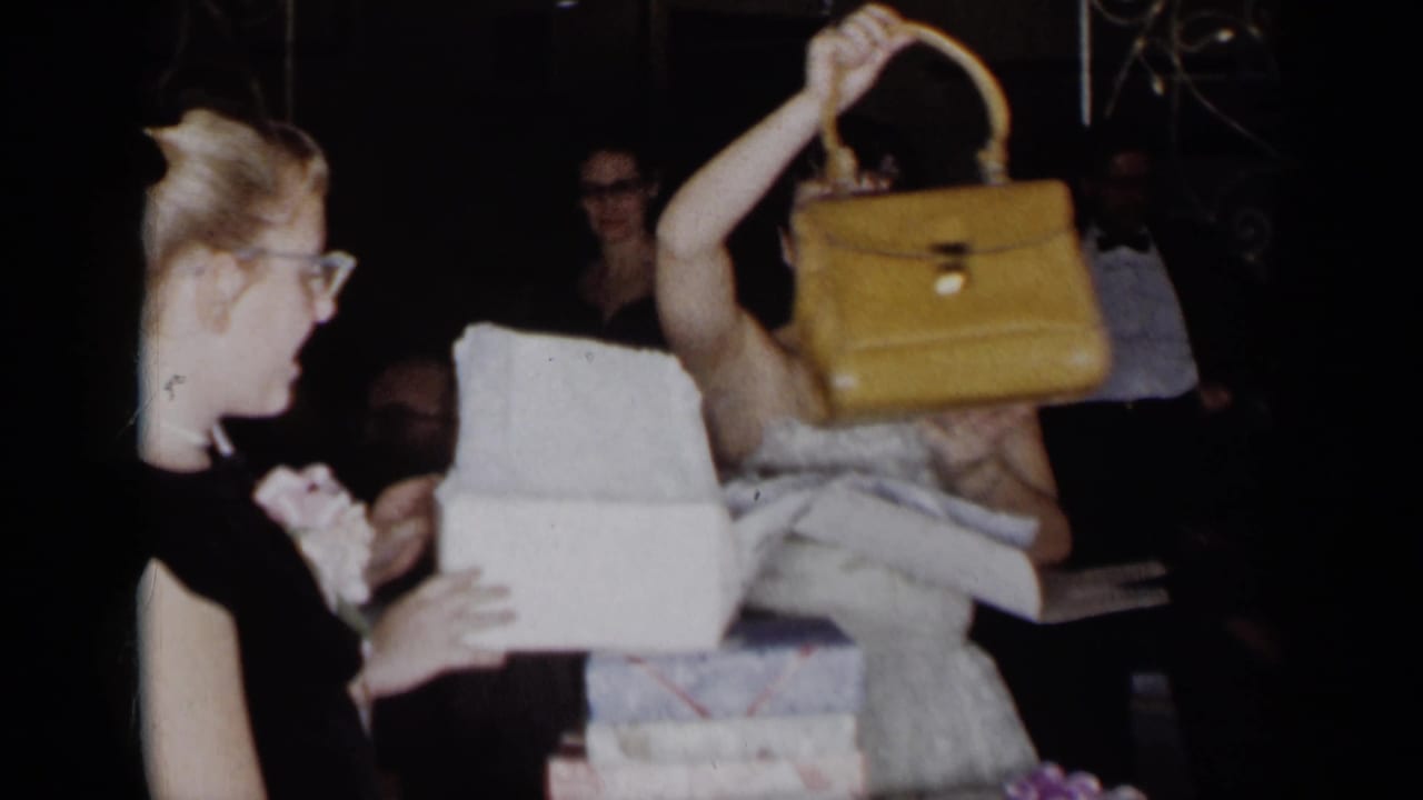 Woman lifts a yellow handbag from a white gift box while another woman stands nearby.