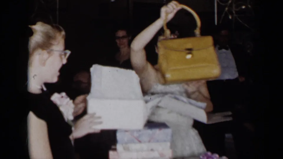 A young woman raises a structured yellow purse from its box during a formal 1954 New York party, with guests and presents around her.