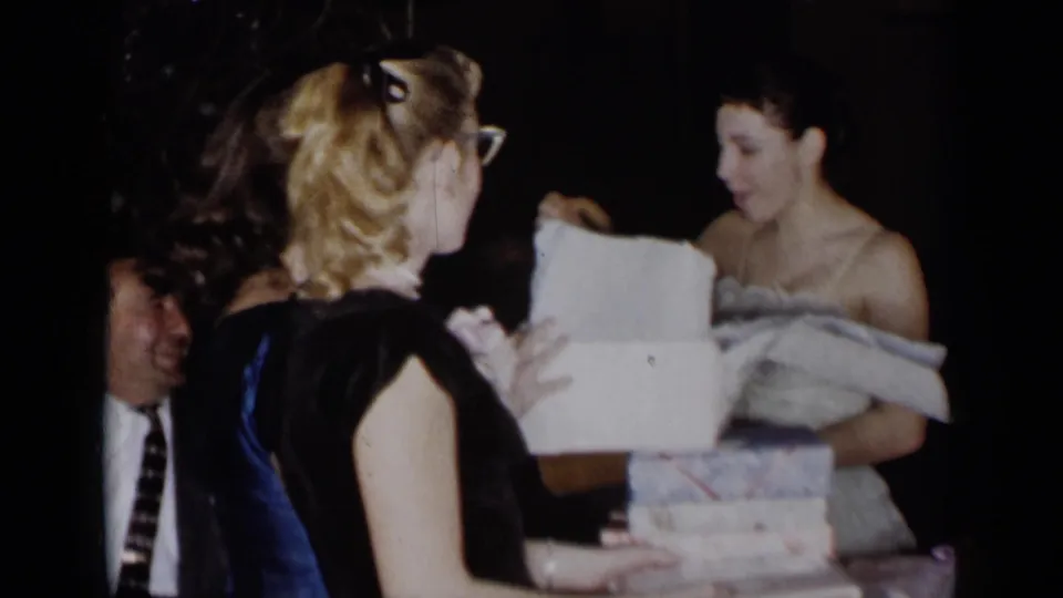At the formal party, the woman stands by the open box and stacked presents after revealing the handbag, with another guest seen from behind.