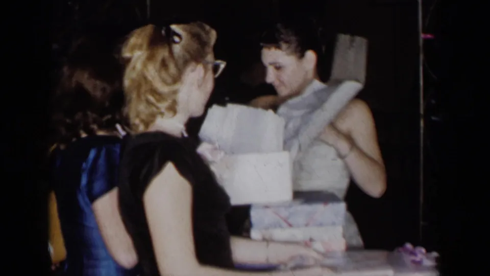 The party scene continues as the recipient holds or adjusts a light wrap near the open gift box, surrounded by formally dressed guests.