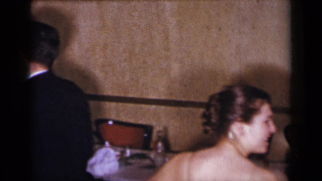 1954 New York wedding reception frame with the bride in a strapless gown near tables while a suited man stands at the left edge.