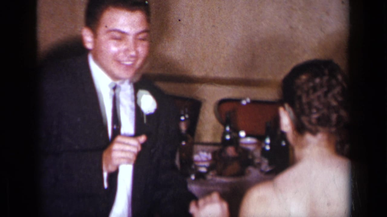 The groom smiles in a dark suit with a white boutonniere as he faces the bride during a 1954 New York wedding reception.
