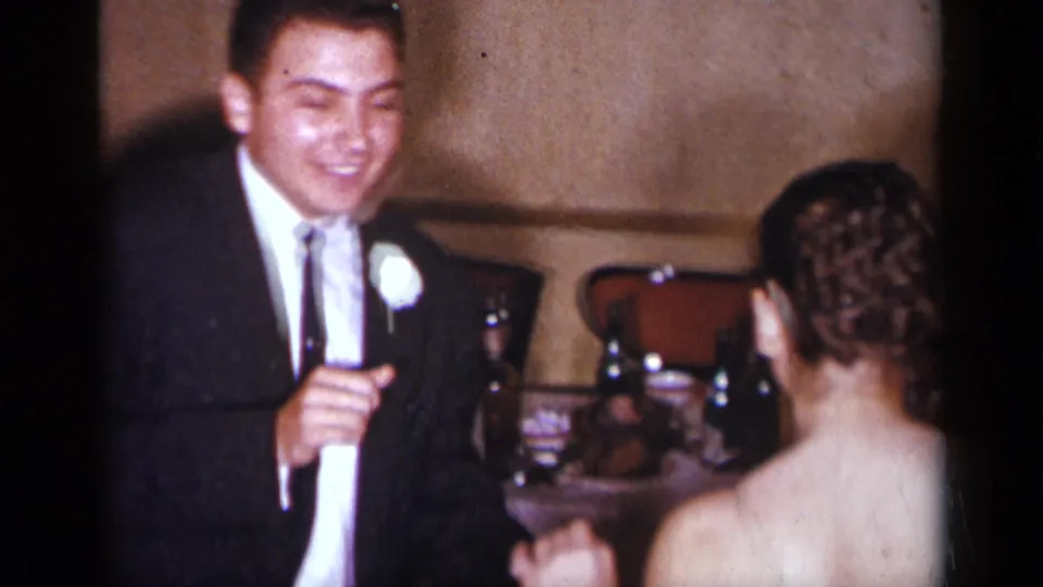 The groom smiles in a dark suit with a white boutonniere as he faces the bride during a 1954 New York wedding reception.