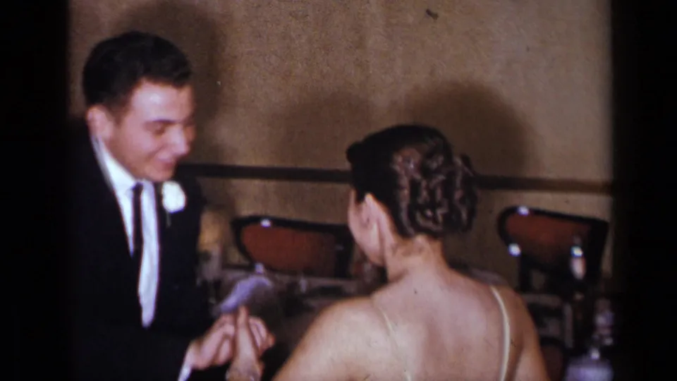 At the New York wedding reception, the groom leans toward the bride and appears to hold her hand beside tables and chairs.
