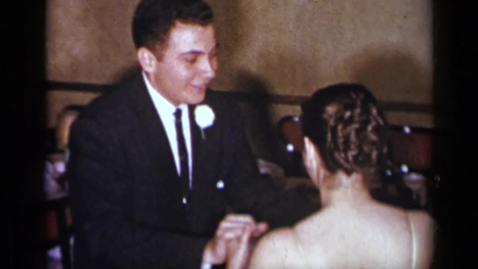 The groom and bride hold hands and stand close in a dim 1954 New York wedding reception interior.
