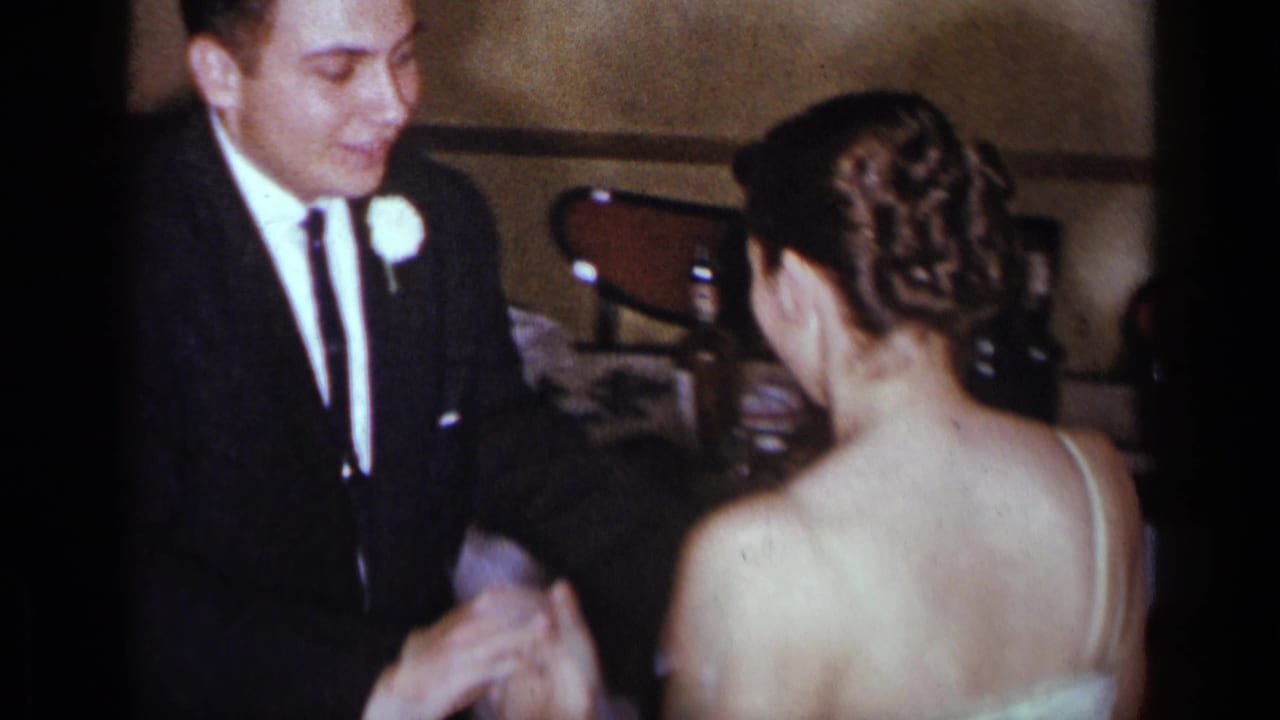 Man in a dark suit stands beside a woman in a white gown, seen from behind indoors.