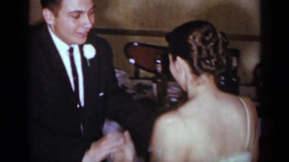 In a 1954 New York home-movie frame, a formally dressed man stands close to a woman in a white gown with her back to the camera.
