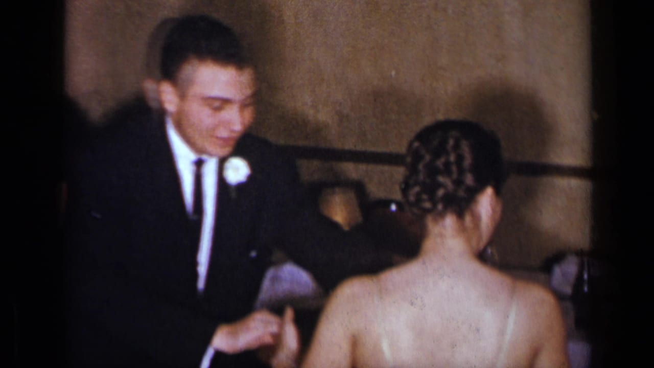 The man in formal wear appears to adjust the woman's dress strap in a dim New York interior documented on 8mm film.