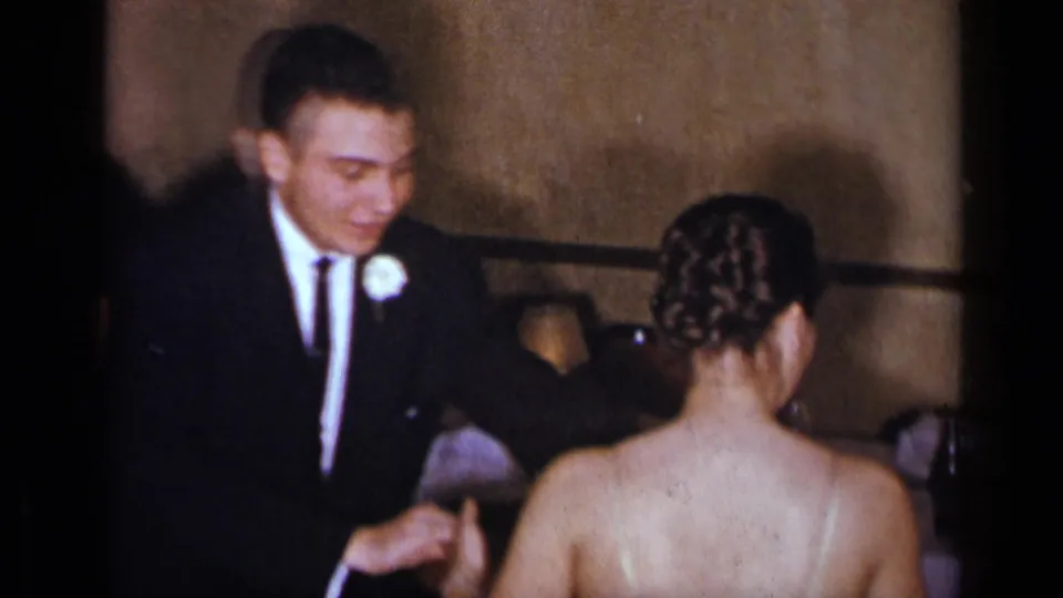 The man in formal wear appears to adjust the woman's dress strap in a dim New York interior documented on 8mm film.