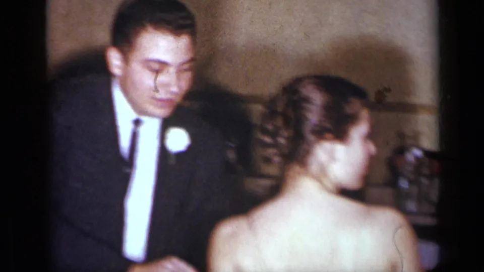 A close 1954 home-movie frame shows the formally dressed couple near a textured wall, with the woman turning partly toward the camera.