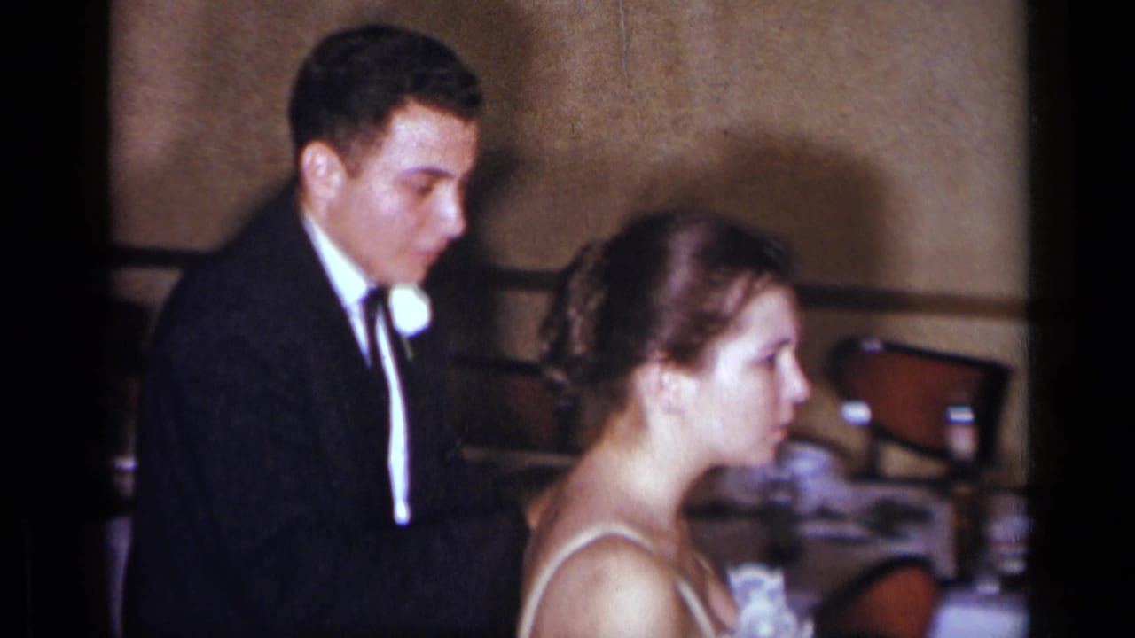 In 1954 New York wedding footage, a young groom in a dark suit and bride in a white gown appear close together in a banquet hall.