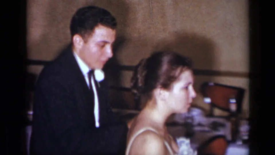 In 1954 New York wedding footage, a young groom in a dark suit and bride in a white gown appear close together in a banquet hall.