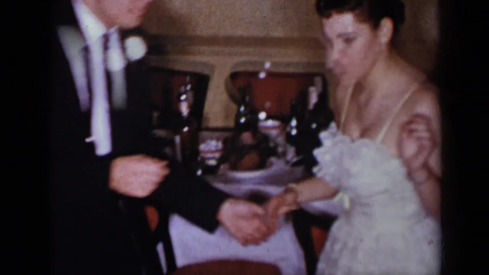 The groom in a dark suit and the bride in a white gown hold hands beside a banquet table in the 1954 New York reception footage.