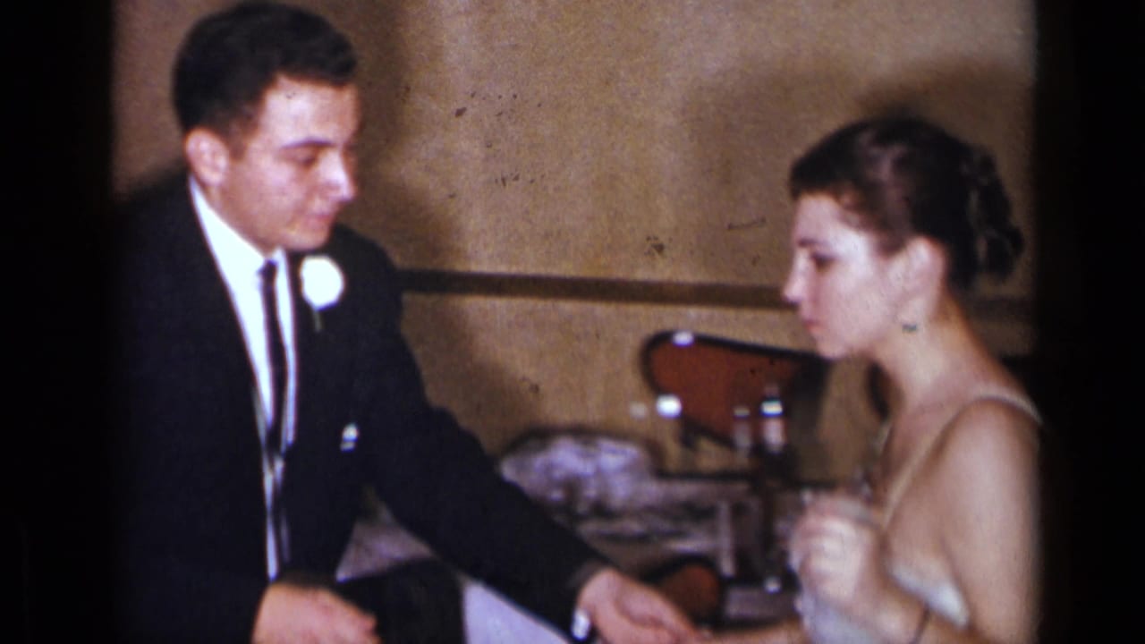 Man in a dark suit with boutonniere faces a woman in a light strapless gown.