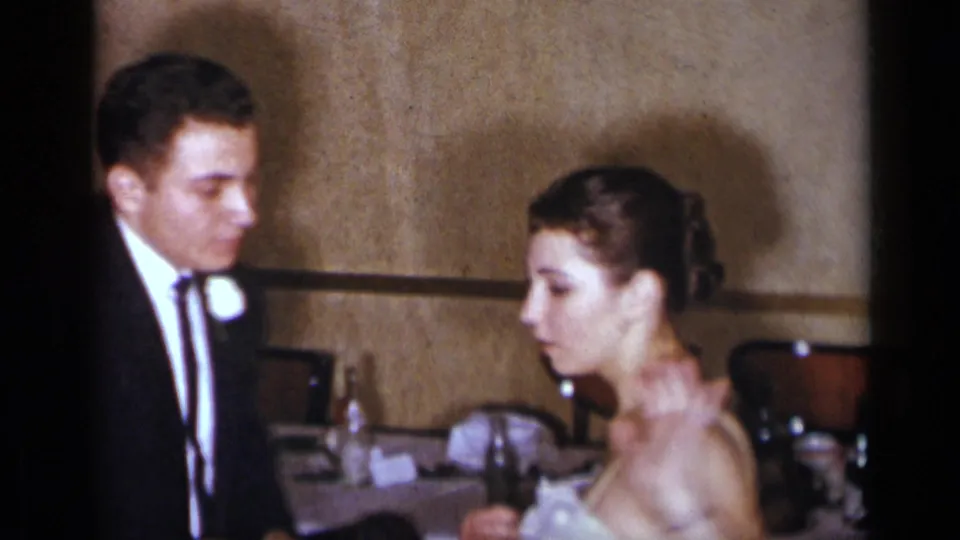A 1954 New York home-movie frame shows a man in a dark suit and a woman in a light gown seated in a private indoor setting.