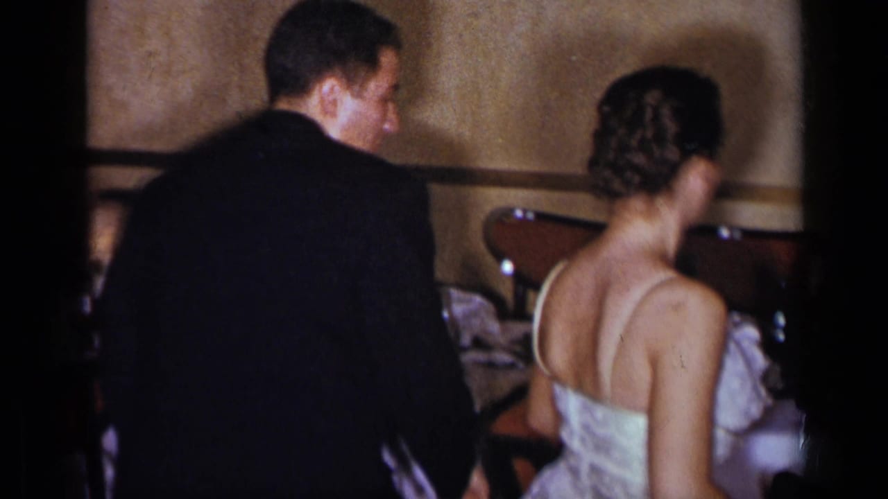 A suited man and woman in a light formal dress stand close together inside a 1954 New York event space.