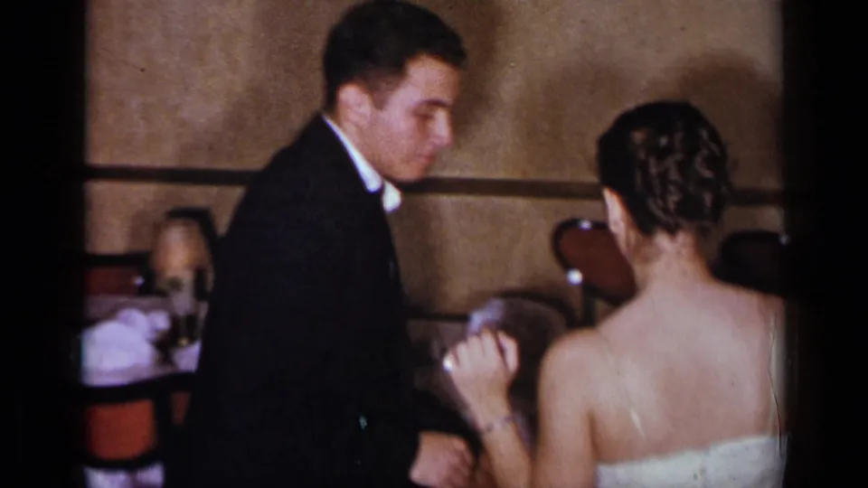 A formally dressed couple faces one another in a dim 1954 New York interior, with banquet chairs visible behind them.