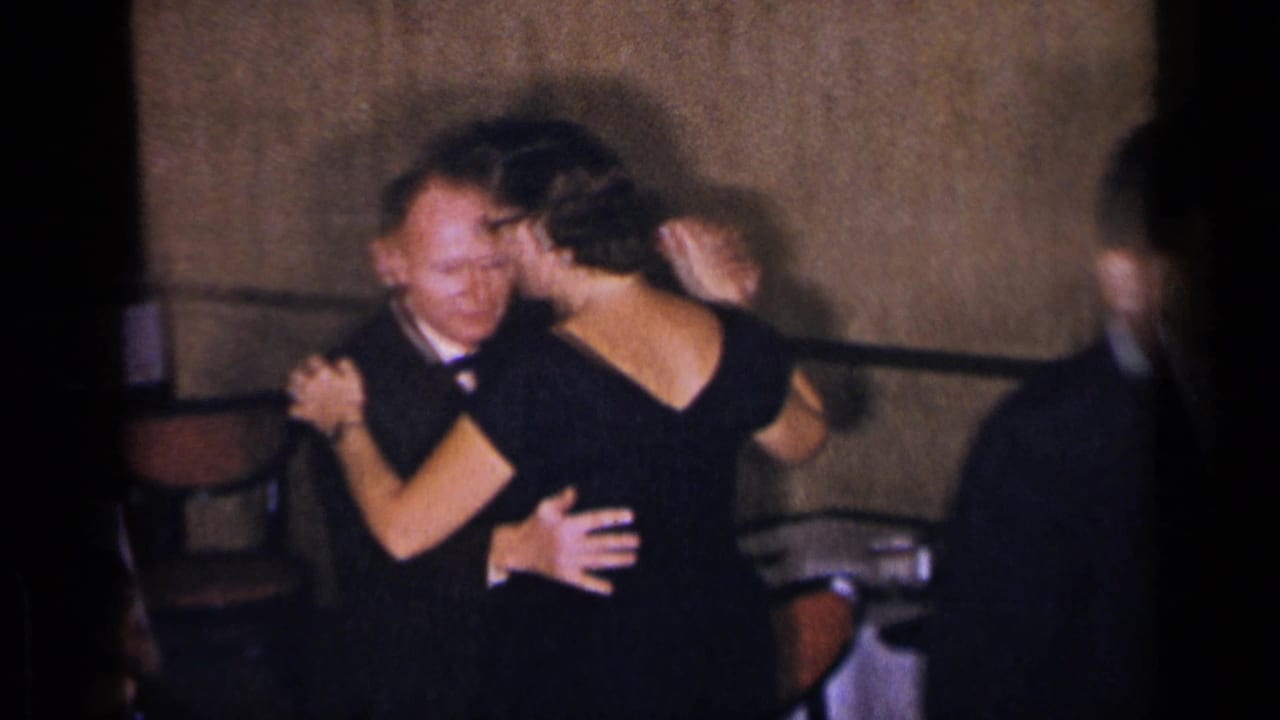 A tuxedoed man and a woman in a black evening gown dance closely in a dim New York restaurant interior, with another guest at the edge.