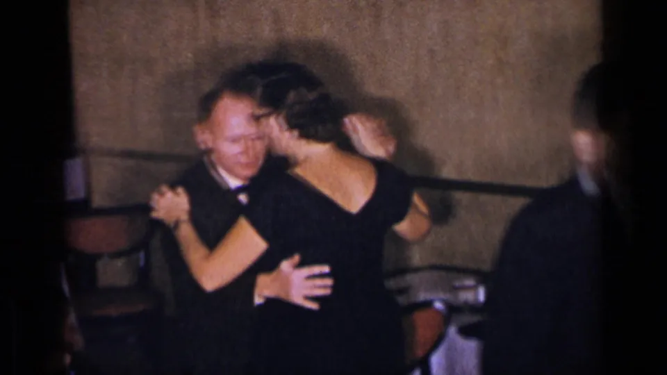 A tuxedoed man and a woman in a black evening gown dance closely in a dim New York restaurant interior, with another guest at the edge.