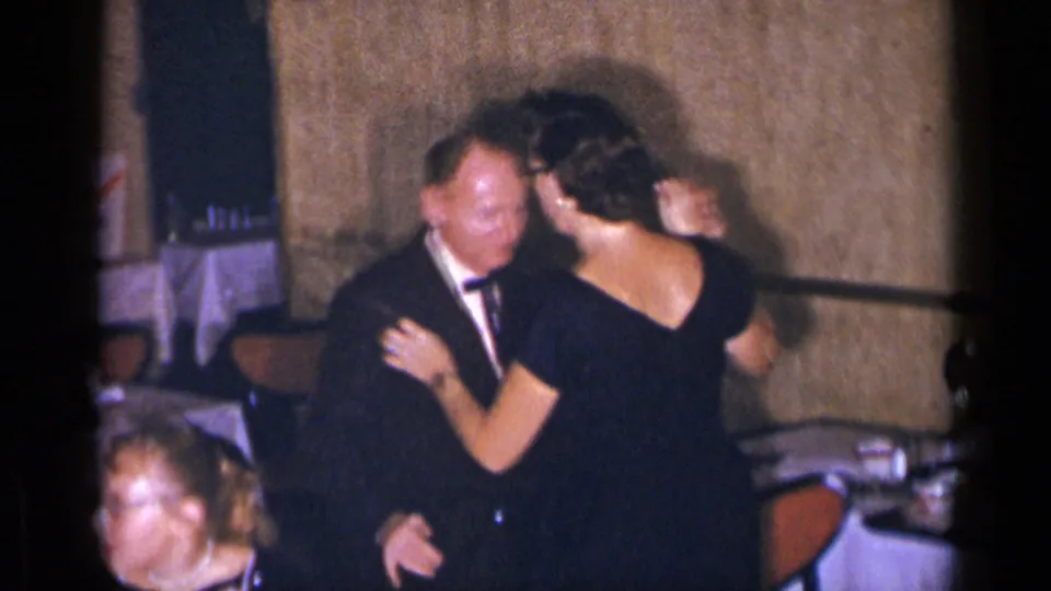 The dancing couple remains close beside restaurant seating, while a seated woman is partially visible in the foreground of the 1954 New York home movie.