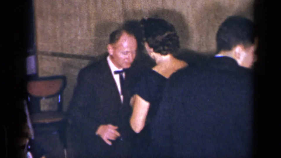 The woman in the black gown turns toward the tuxedoed man in the restaurant, partly obscured by another guest moving through the frame.