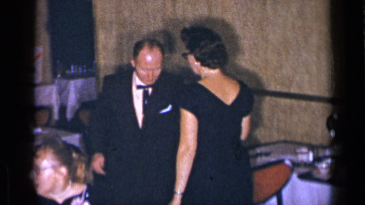 Man in tuxedo stands beside a woman in a black dress in a formal interior.