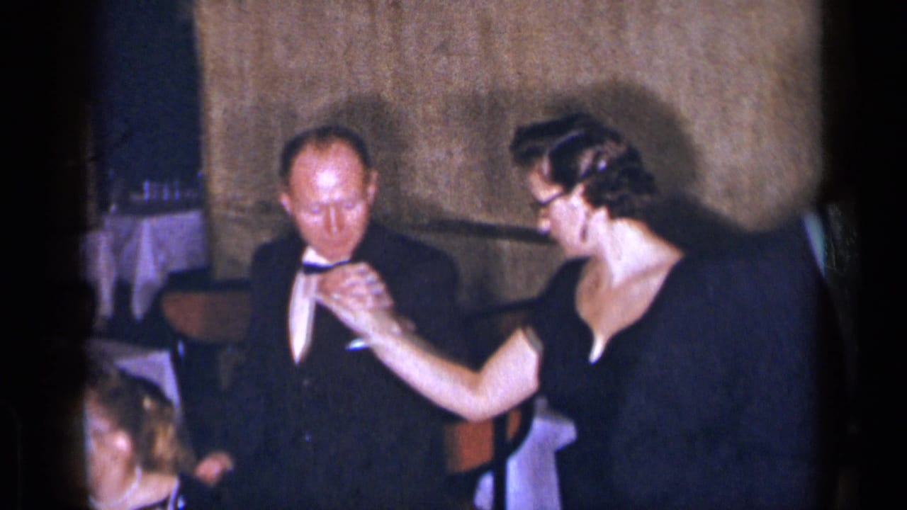 1954 New York home-movie frame of a woman in a black dress reaching toward a man's bow tie in a formal interior.