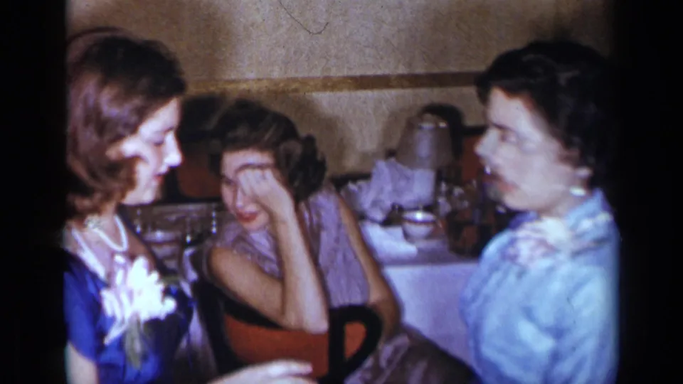 1954 New York home-movie frame showing women in mid-century dresses seated at a table in a softly lit interior.