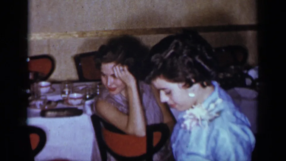 1954 New York home-movie frame of two women seated at a formal table in a private indoor social setting.