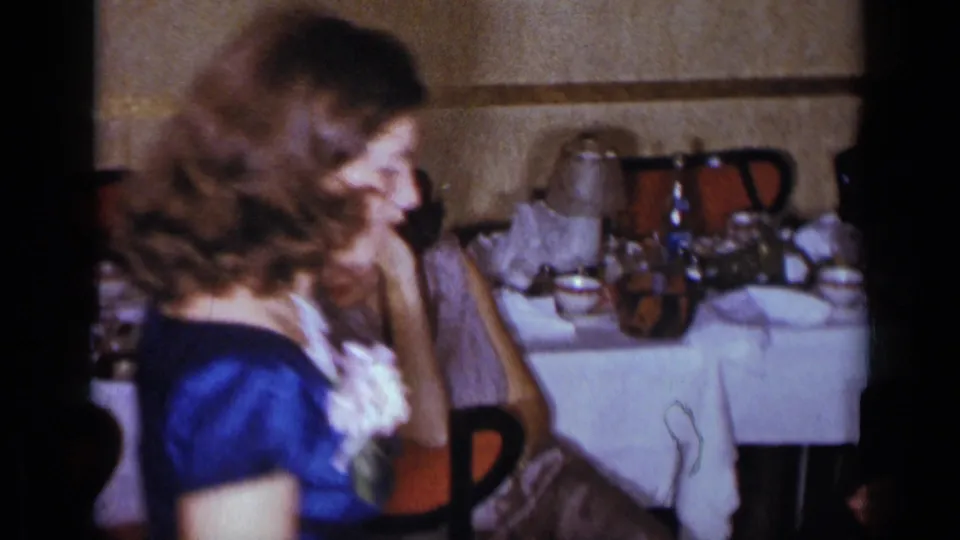 1954 New York frame with a woman in a blue satin dress and corsage moving past another woman seated at a formal dinner table.