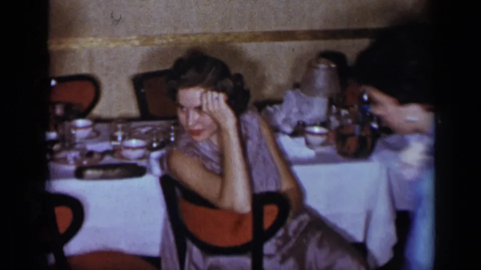 A 1954 New York dinner scene shows a woman in a lavender dress seated beside white linens and glassware as another guest leans into frame.