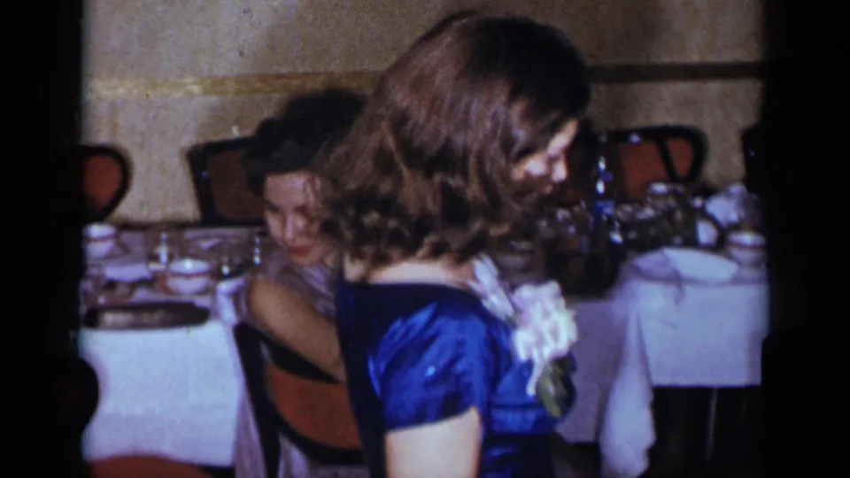 1954 New York home-movie frame of a blue satin dress and corsage in the foreground, with a seated woman and formal table settings behind.