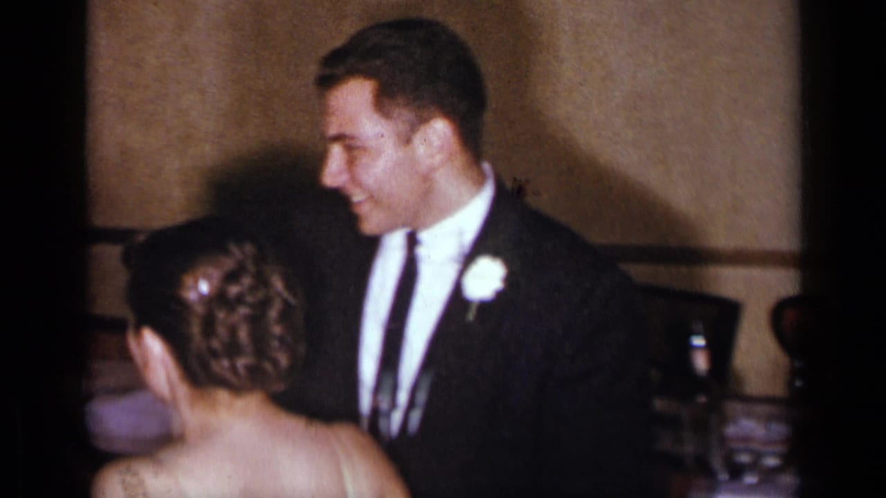 Groom in a dark suit smiles toward the bride, seen from behind.