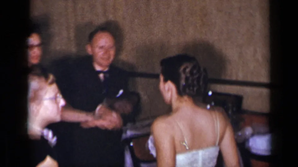 1954 New York home-movie frame showing the bride near older formal guests, with one woman reaching toward her in a banquet room.