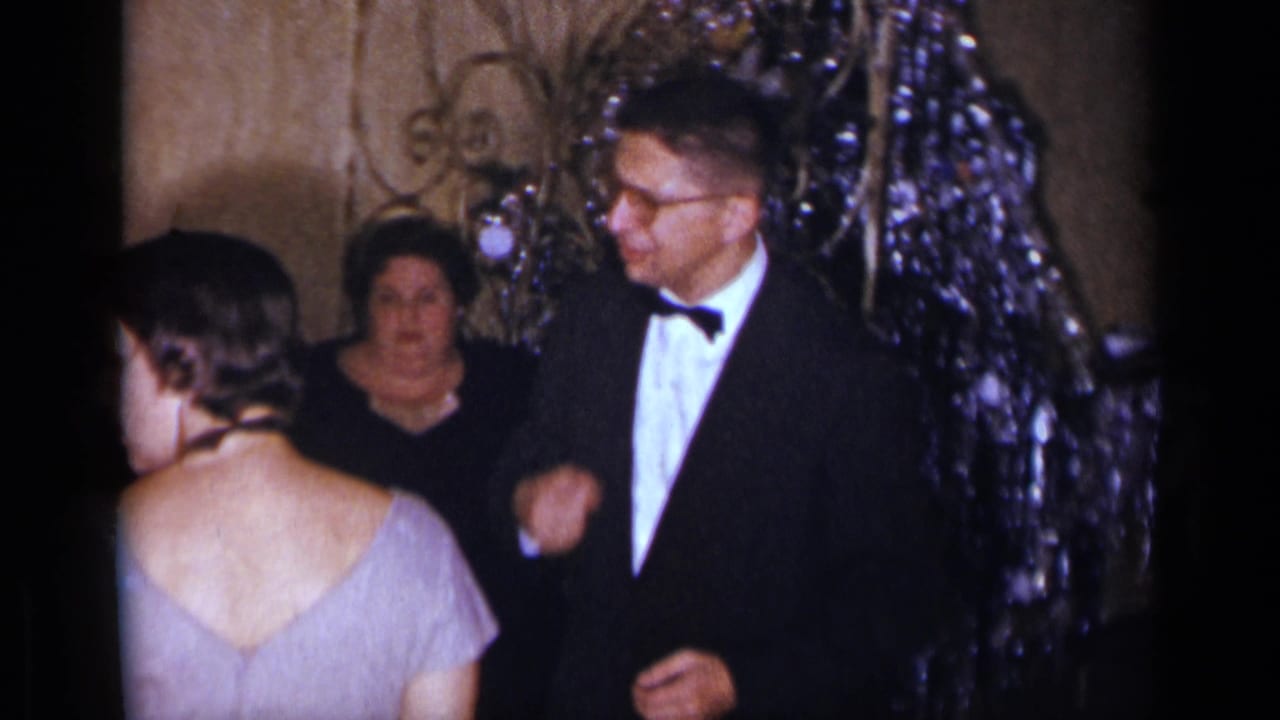 1954 New York party frame with a tuxedoed man and woman in a light dress near shimmering holiday decorations.