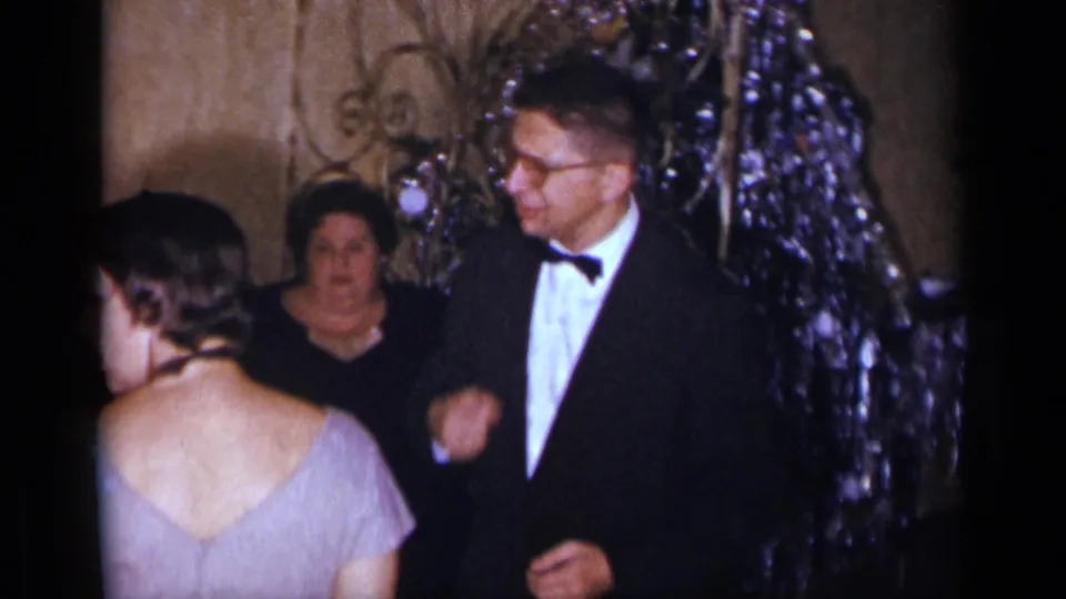 1954 New York party frame with a tuxedoed man and woman in a light dress near shimmering holiday decorations.
