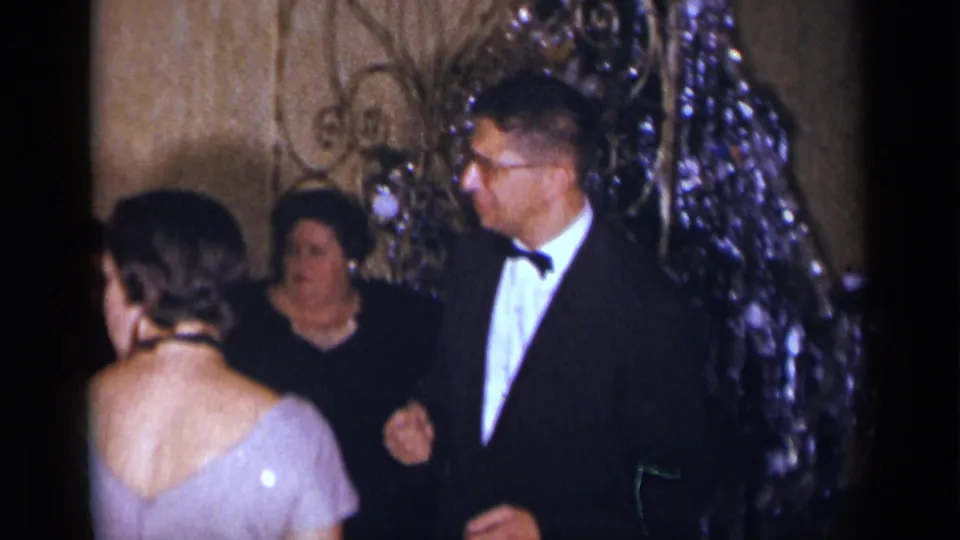 1954 New York home-movie frame showing formal party guests near a reflective holiday backdrop in a private room.