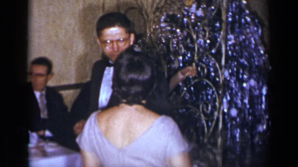 1954 New York party frame with a tuxedoed man, a woman in a light dress, and shiny decorations in a private interior.