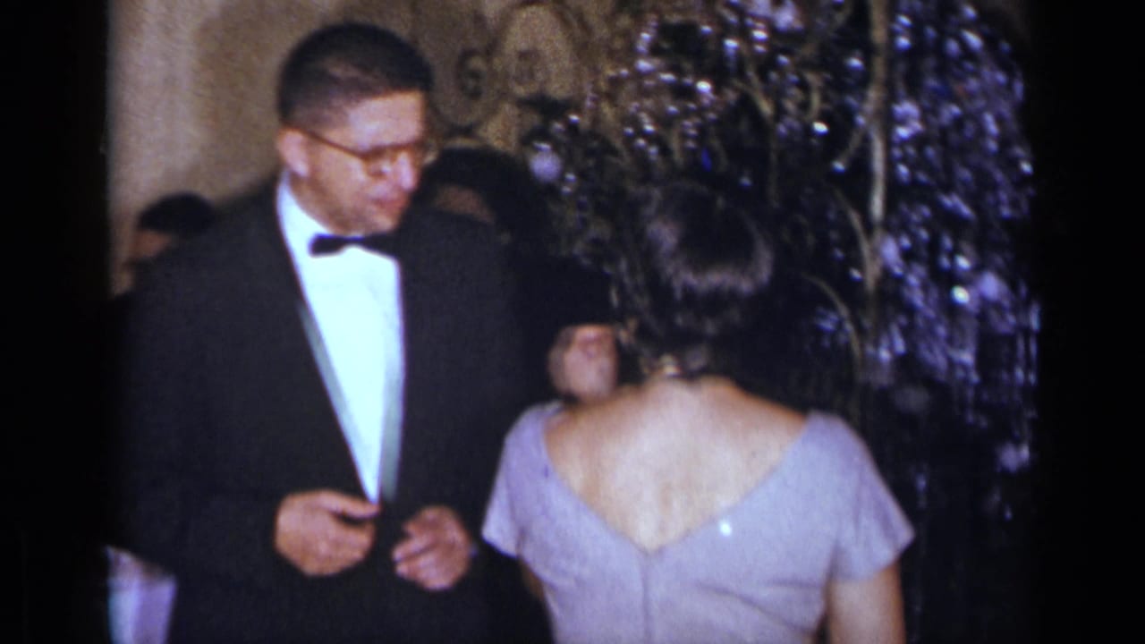 Man in a tuxedo faces a woman in a pale dress at an indoor formal event.