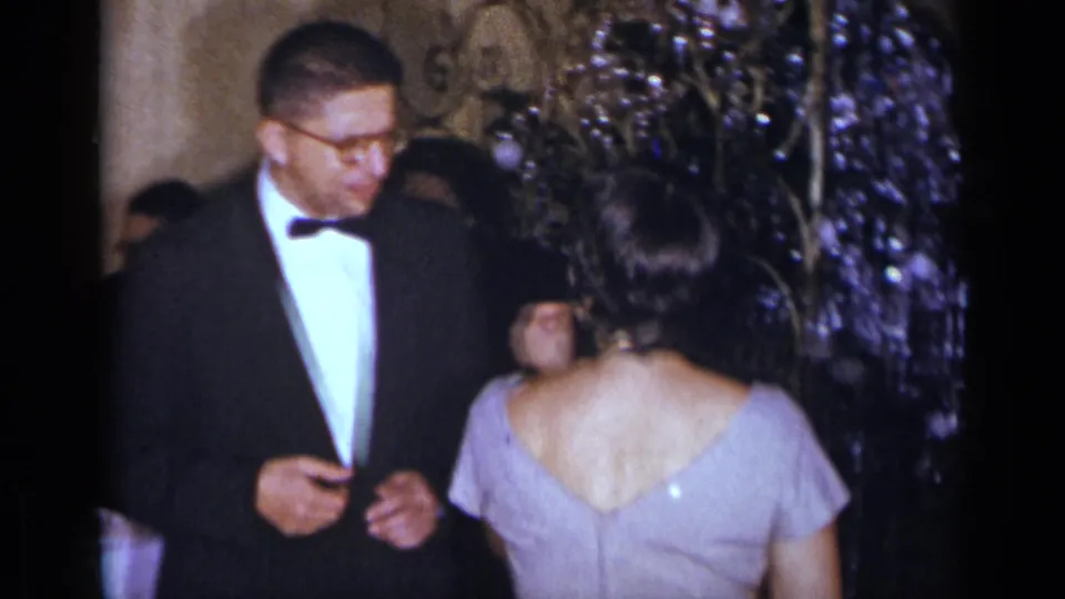 At a 1954 New York formal gathering, a tuxedoed man stands near a woman in a pale purple dress as guests sit behind them.