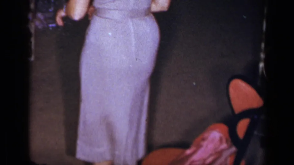 The woman in a backless pale purple dress is seen from behind during the 1954 New York indoor reception, with chairs and guests nearby.