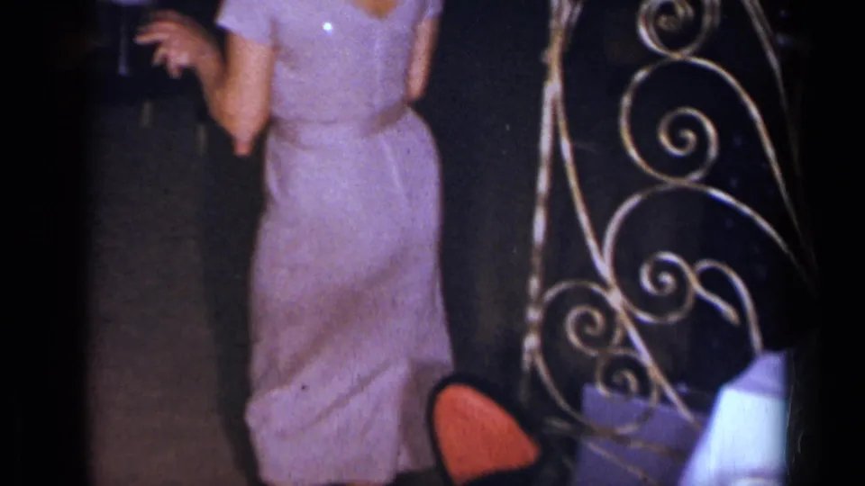1954 New York home-movie frame showing a woman in a sparkly purple dress dancing near decorative metal scrollwork.