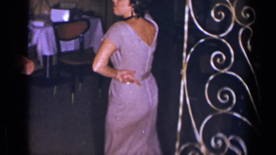 The woman turns with one hand at her back, with chairs, a small table, and decorative scrollwork visible in the party room.