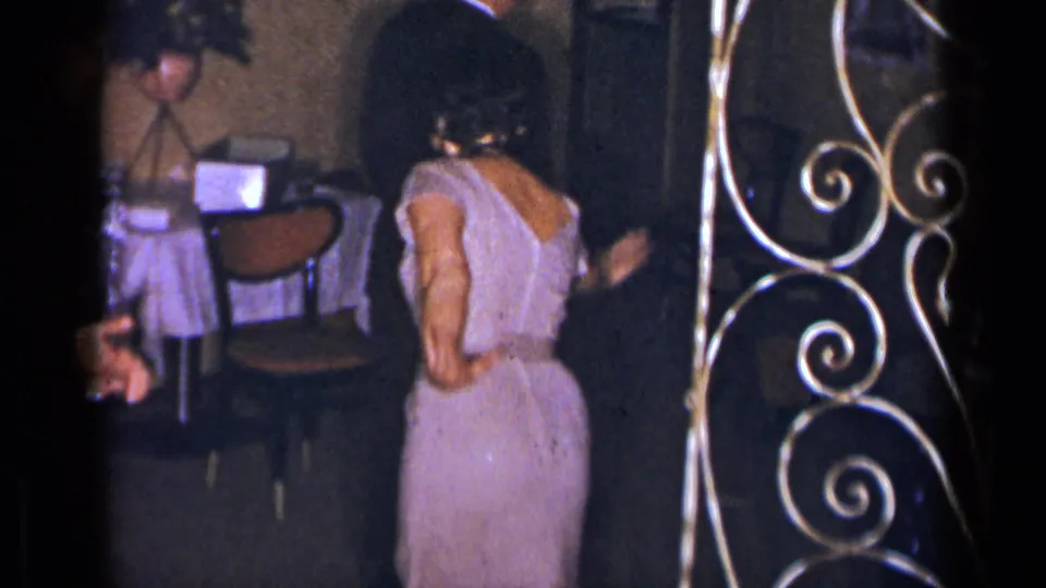 In the dim 1954 New York party setting, the woman stands near a table, chairs, and ornate metalwork while turning away.
