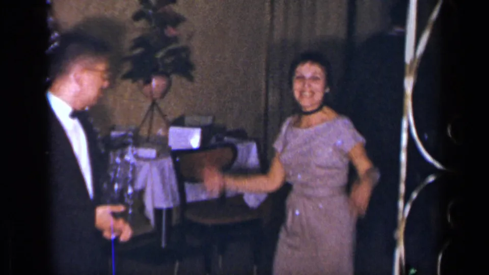A woman in a sparkly purple dress smiles and gestures beside a tuxedoed man, with table furnishings and a plant behind them.