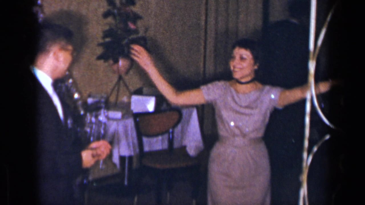 Woman in a pale purple dress dancing beside a tuxedoed man in a dim party room.