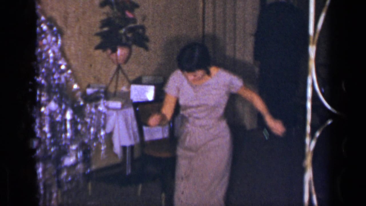 1954 New York home-movie frame of a woman in a pale purple dress dancing near tables, plants, and silver party decor.