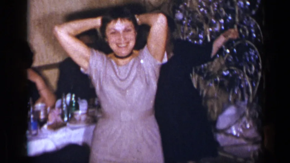 1954 New York home-movie frame showing the dancing woman smiling with hands behind her head beside banquet tables and guests.