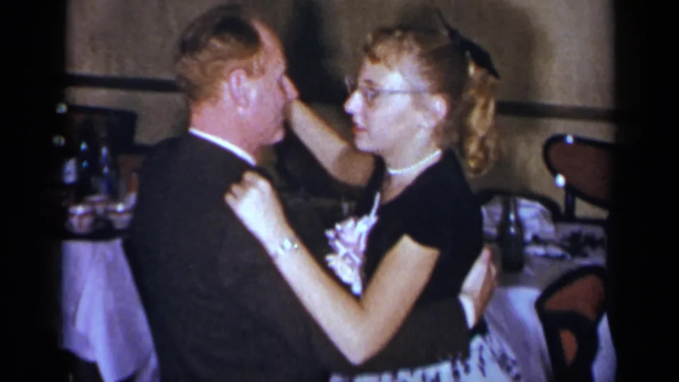 In a 1954 New York home-movie frame, a suited man and a woman with glasses, pearls and a hair bow dance closely beside set tables.