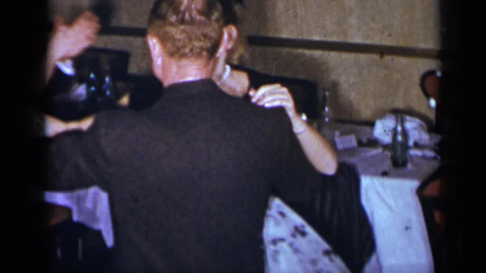 The camera catches the dancing couple from behind in the New York interior, with table settings and bottles visible around them.